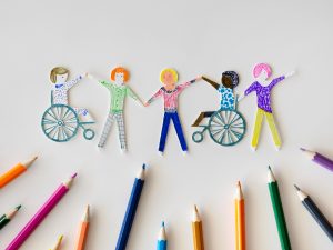 Artsy colorful paper cut outs of 5 students of diverstiy, two using wheelchairs. Color pencils pointing at the group. 